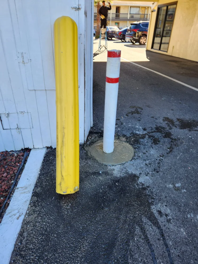 New white steel bollard with red reflective tape set in fresh concrete, next to a yellow plastic bollard cover