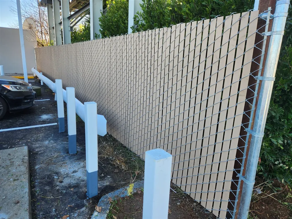 Chain link fence with tan privacy slats screening a parking area, with painted bollards along the curb, built by Palmer Fence