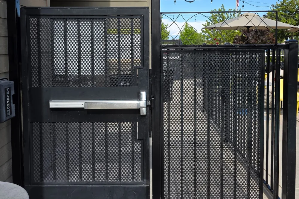 Black expanded metal man gate with a push-bar panic device at a commercial patio, built by Palmer Fence