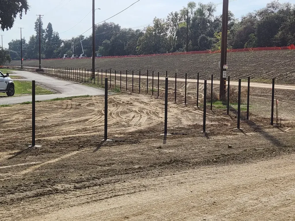 Row of black steel fence posts set in concrete footings along a dirt lot, before the panels are hung