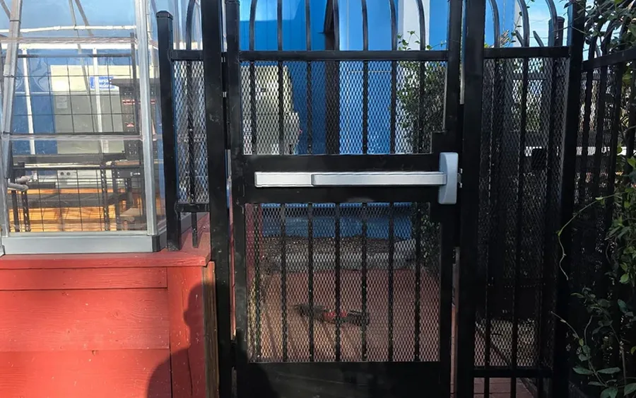 Black curved-top steel gate with a panic bar and expanded metal beside a greenhouse